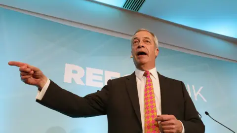 PA Media Nigel Farage, wearing a black suit with a white shirt and a fuschia pink and gold houndstooth tie, points during a speech.