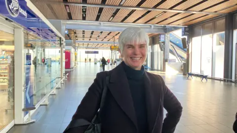 Charlotte with short grey hair and a black top and coat, standing smiling outside Boots in Reading station with signs for platforms 10 and 11 behind her