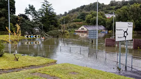 Athena Picture Agency A flooded roundabout with a fallen tree in the background. A road sign partially by the water can be seen on the right of the image.