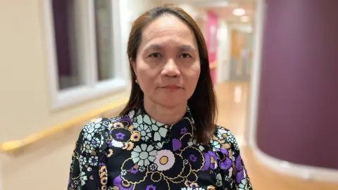 Arlene Elano has medium length brown hair. She is wearing a black blouse with purple, pink and light blue flowers.