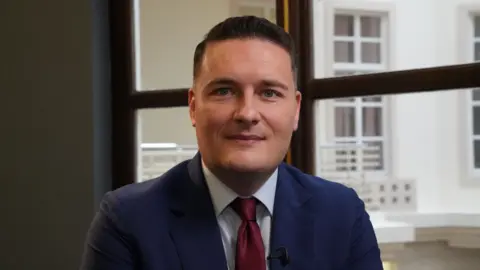 John Fairhall/BBC Wes Streeting has short dark hair and is wearing a navy suit jacket, whie shirt and dark red tie. He is smiling in front of a window indoors.