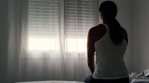 Woman sitting on a bed staring out of a window covered by a net curtain