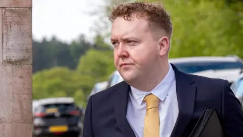 PA Media Jordan Linden arriving at court. He has short brown hair and is wearing a dark blue suit, pink shirt and yellow tie.