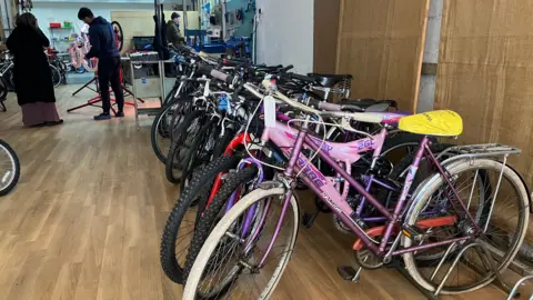 Grace Wood/BBC Bikes lined up against a wall. In the background a man and woman fix a bike in a workshop.