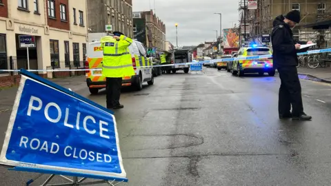 Police officers stand on North Street in Bristol. In the foreground is a blue and white "road closed" with white text on a blue background