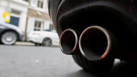 An exhaust pipe on the back of a car. A wheel can be seen, with two cars out of focus in the background
