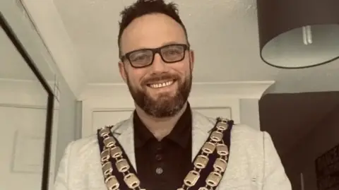 James Clayton A man with glasses, dark hair and a beard in a white suit wearing ceremonial chains of office in a room with white walls