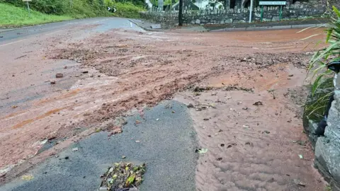 Rachel Jagger A flooded road mixed with mud besides a pub in Totnes. The water is brown with mud and there is a brick wall on the opposite side of the junction.