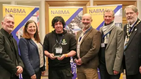 South Molton Town Council Representatives from the town and the market stood in a line. One in the middle is holding the glass award. 