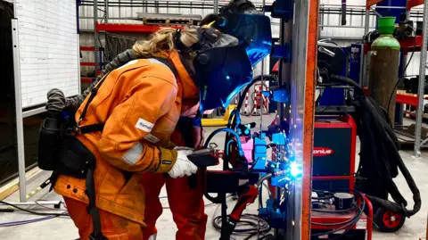 Two welders in full PPE with darkened visors stand in front of a sheet of steel. Blue sparks fly from where the welder is striking the metal.