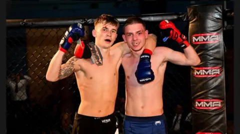 Two young men stand in a Mixed Martial Arts cage and grin at the camera. One has his arm around the other. They both wear boxing gloves.