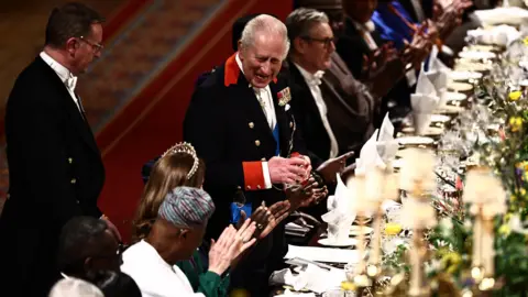 PA Media King Charles III in a black suit jacket and trousers and white waistcoat, smiling and standing while addressing the banquet at a long table.