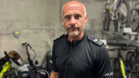 Un bărbat chel, cu barbă scurtă, privește direct în cameră. Poartă un top negru cu mânecă scurtă, pe care scrie „poliție” pe mâneca stângă. În spatele lui se află biciclete lipite de un perete și pe un suport.
