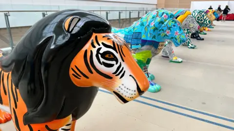 A row of the brightly coloured lions inside in a large warehouse. The one closest to the camera is covered with a tiger print, while the others are all multicoloured.