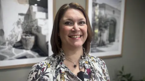 A female teacher, who is the director of music at the school, wearing a floral top and smiling to camera