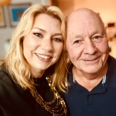 Nicky West A blonde-haired woman and a bald older man have their heads touching as they smile directly at the camera. The lady wears a black blouse with a layered gold chain necklace. The gentleman wears a blue polo shirt unbuttoned at the top and has a small white detailing on the collar. The background behind the pair is blurred out and conceals their surroundings.
