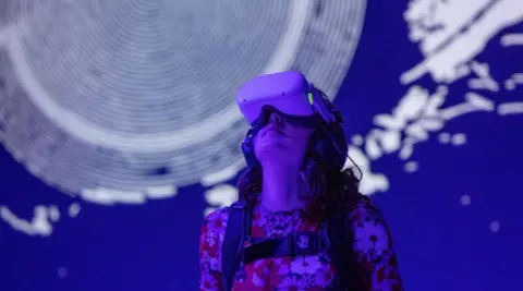 Woman wearing a VR headset at the exhibition, gazing upwards in front of a blue and white exhibition display