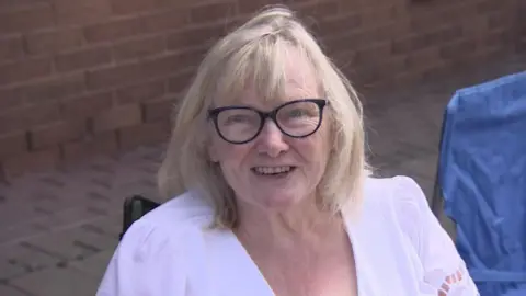 Carol está sentada en una silla al borde de la calle. Lleva gafas, una de ellas blanca.