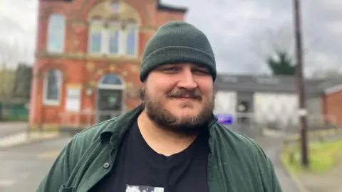A man with a beard wears a green hat. He stands in front of a brick church.