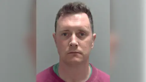 Norfolk Police A custody photograph of Simon Taylor who is looking directly at the camera with a neutral expression. He is wearing a green T-shirt with a pink pullover, over the top.