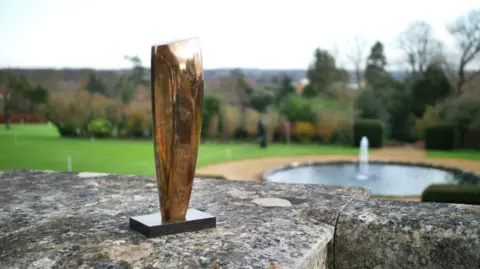 A plain polished bronze sculpture on a stand. It appears to be the size of a forearm and has been placed on a stone-capped wall in the open air. In the background is a lawn with a round pond and fountain.