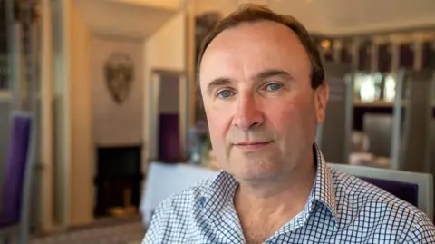 A man with short, brown hair looks at the camera. He is wearing a blue and white checked shirt and appears to be sitting in a restaurant 