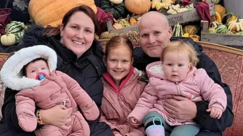 Submitted Mike and Laura Barlow, both wearing black coats, hold three young girls, all dressed in pink, at a Halloween event.