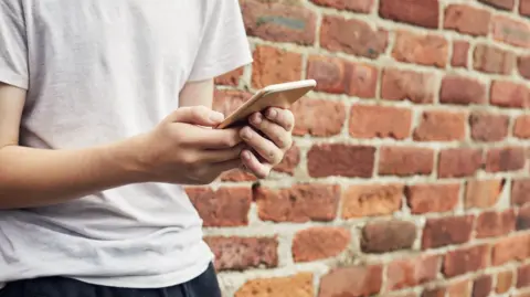 A teenage boy standing against a brick wall types on his smartphone.
