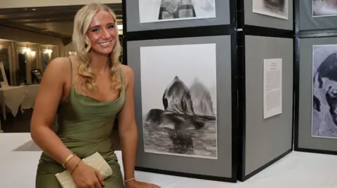 Emma Price, tanned and smiling, with long blonde hair in loose curls. She is wearing a strappy green satin dress and holding a cream beaded clutch bag. Next to her is a stand of black and white sketches.