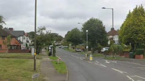 A road in a residential area with houses, trees and grass verges on either side and a crossing point marked with bollards