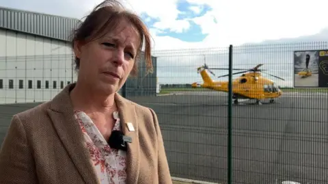 Karen Jobling is wearing a beige coat with a floral blouse. Her head is tilted to the right. She has brown hair. Behind her is a yellow helicopter.