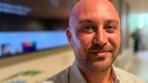 Phil Shepka/BBC Ricky Ioannides stares at the camera. He wears a white shirt with an orange lanyard. 
