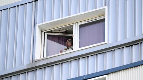 PA Media A person standing at an open window of a flat, wearing a hoodie and a white jacket. There is light blue cladding on the outside of the building, and the window has a white frame.