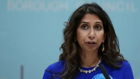 Suella Braverman talks into a microphone while wearing a royal blue top, a blue rosette, and a large necklace.