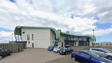 Google The Tuned In! centre. It is a white building with a green roof. Several cars are parked outside the building.