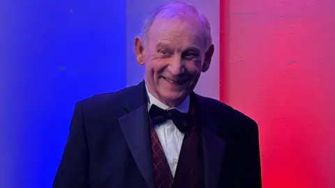 Elly Fiorentini/BBC An older man wearing a tuxedo smiles, looking off to the side of the camera. The wall behind is lit red, white and blue.