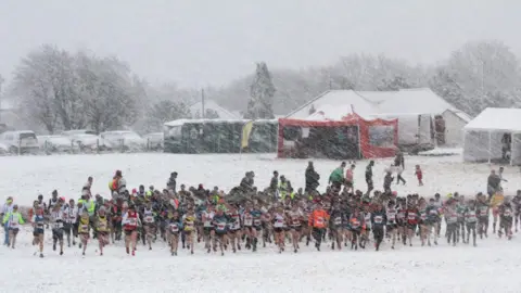 Ian Dunn A crowd of people running in the distance on a field covered in snow. Snow is coming down heavily around them. The cars parked in the distance are completely covered. There are tents and portaloos at the back of the field.