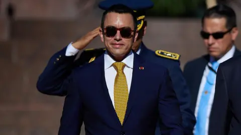 BBC Ecuadorean President Daniel Noboa wears a blue suit, yellow tie and sunglasses. Someone in military uniform salutes behind him as another man in a suit, pale blue tie and sunglasses stands, blurred out, behind him.