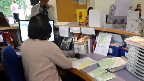 A GP surgery reception. The receptionist has her back to the camera and is talking to a female patient who is standing at the desk. The desk has a phone, PC, prescriptions and filing systems. Beyond is a waiting room where two women are sitting.