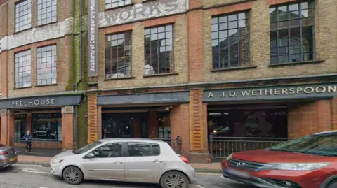 An exterior view of the Wetherspoon pub in Bridge Street, Guildford, a red and brown brick building, with a line of cars waiting at traffic lights in front of it.