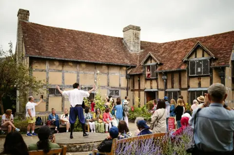 Sam Allard / Shakespeare Birthplace Trust Actors perform in the garden of Shakespeare's Birthplace. A man is standing on a raised area with his arms held up in a dramatic gesture and people are sitting on benches around him watching. Shrubs and plants from the garden can be seen, and the half-timbered, 16th Century Tudor building, with its wooden framework and wattle and daub infill, is in the background.