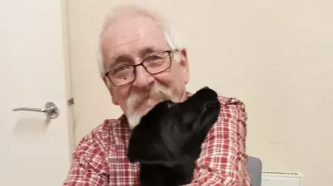 A man with grey hair and glasses wearing a red and white checked shirt and cradling a black dog in his arms.