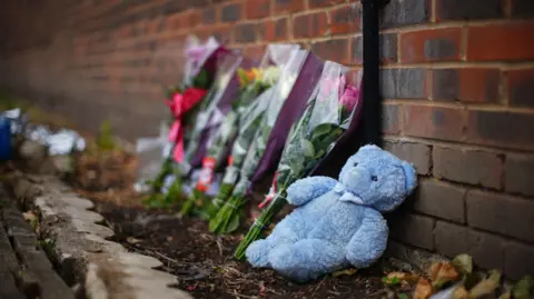 An image taken from the ground, a teddy bear and floral tributes lie against a brick wall