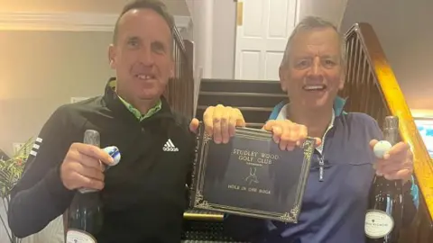 Studley Wood Golf Club Steve Wilmhurst and Liam Nairn smile and hold up the Studley Wood Golf Club hole-in-one book. They each have a bottle of champagne and a golf ball.