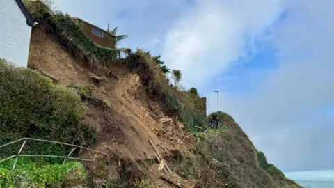 Part of a cliff seen collapsed on the side of the hill, a small building sits at the top and two people are seen in the distance on a path.