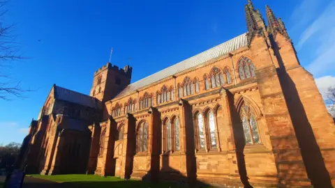 BBC Weather Watchers/Gadabout A general view of Carlisle Cathedral taken from its side. The building in red sandstone has arched windows all along its side, with gothic-type detailing and turrets. The sky is blue and bright.