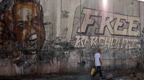File photo showing a man walking past a portrait of Marwan Barghouti (L) on a section Israel's West Bank separation barrier, near the Qalandia checkpoint, in the occupied West Bank (22 September 2025)
