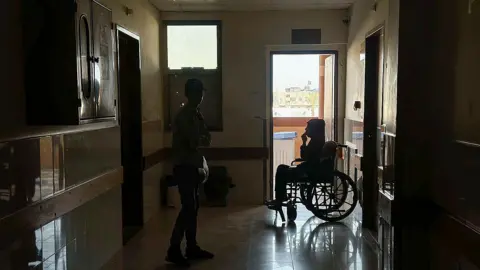 AFP A person stands near a patient sitting in a wheelchair in a dark corridor during a power outage at Nasser hospital in Khan Younis, southern Gaza (20 June 2026)