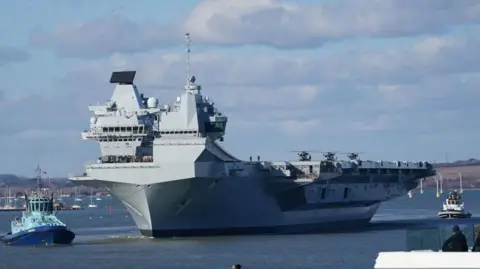 PA Media HMS Prince of Wales, an enormous, grey vessel with three helicopters visible on its flight deck, is towed along the Mersey Estuary by a blue tug boat