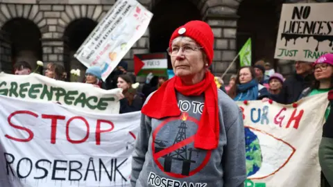 Getty Images Protesters outside Edinburgh's Court of Session with white banners saying "stop Rosebank". In the foreground is a woman with glasses, a red hat and scarf, and a grey jumper with an anti-Rosebank slogan
